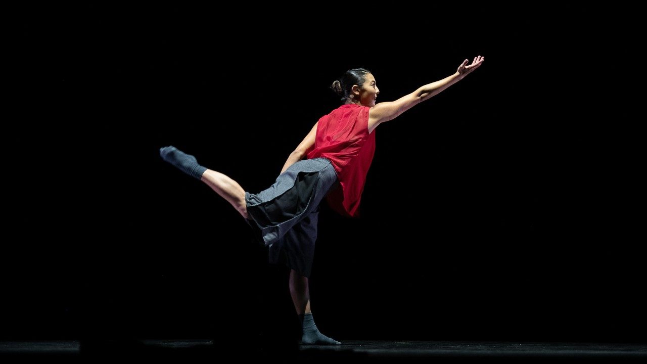  A dancer from Ballet Hispanico dances on stage. She stands on one foot, the other extended behind her, bent at 90-degree angle at the knee, with one arm extended in front of her.