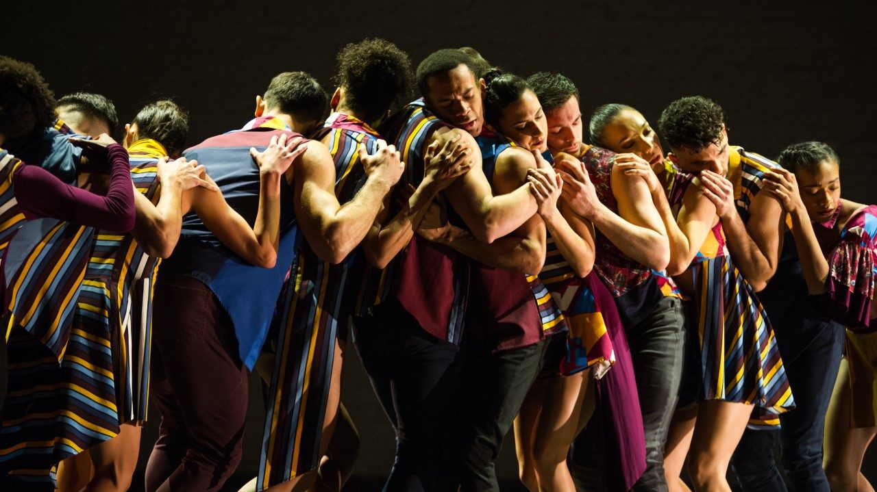  The dancers of Ballet Hispánico on stage in multicolored costumes huddle in a group and rest their heads on each other's shoulders.